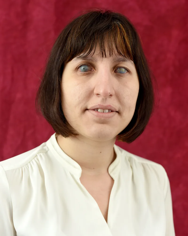 Elena Karamanska, head and shoulders, with a dark brown bob and fringe, in a cream blouse against a deep red backdrop, smiling slightly.