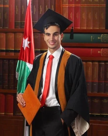 Hasan Tayem smiling at his graduation, in a black gown with orange trim, a mortarboard and a red tie, holding an orange folder beside the flag of Jordan, in front of shelves of leather-bound books.